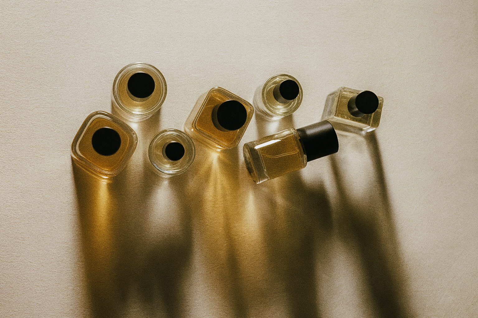 Top-down view of multiple glass perfume bottles arranged vertically on a textured off-white background, lit from the left with dramatic golden shadows and soft reflections. Each bottle is minimalist with a black cap and filled with amber or clear liquid, captured in a vintage, film-style aesthetic.